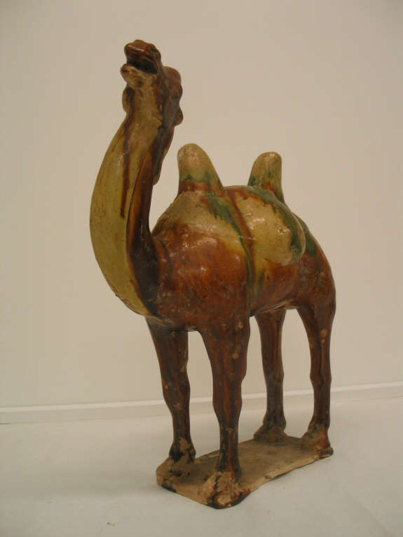 Angled front view of a glossy green, tan, and brown figurine of a camel with a long neck and two humps standing on a short attached rectangular platform.