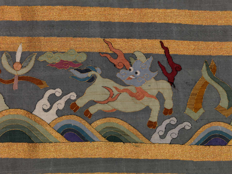 Border detail of the blue-gray robe’s back featuring a green animal leaping over waves, wispy pastel clouds and vegetal motifs encased in gold and blue stripe borders.