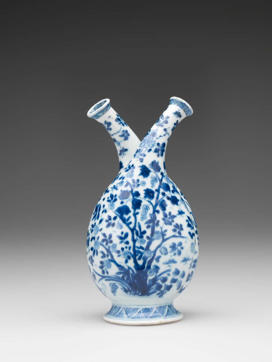 White bottle with two necks, patterned with dark blue leaves. Rounded at the bottom, the form tapers into two elegant spouts, which curve away from each other.