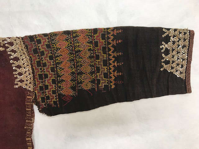 Detail view of a black sleeve on a light background adorned with embroidered orange and black geometric patterns across the top. Along the cuff and shoulder are dense sequin detailing.
