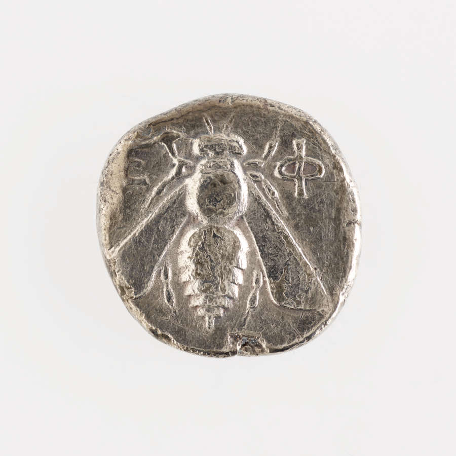 Rough-edged round silver coin embossed with an image of a bee with Greek letters beside it.