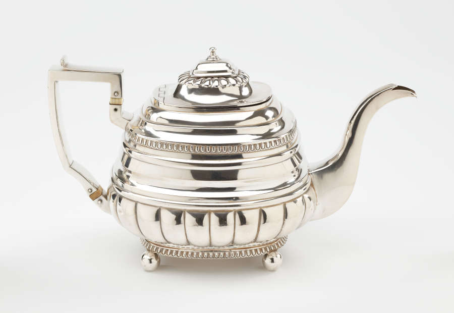 A silver teapot with a decorative angular handle, a rounded square body and spout with sculptural decorations.