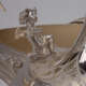 Detail of silver cherub with wings gazing upwards with outstretched arms and wavy hair, kneeling with one knee up at the base of a curved silver surface with floral design. 
