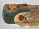 Side view of a coffin with a gilded face, blue-black headdress, inlaid eyes, and a sculpted beard. A red sun above a black and white falcon sits on the shoulder.