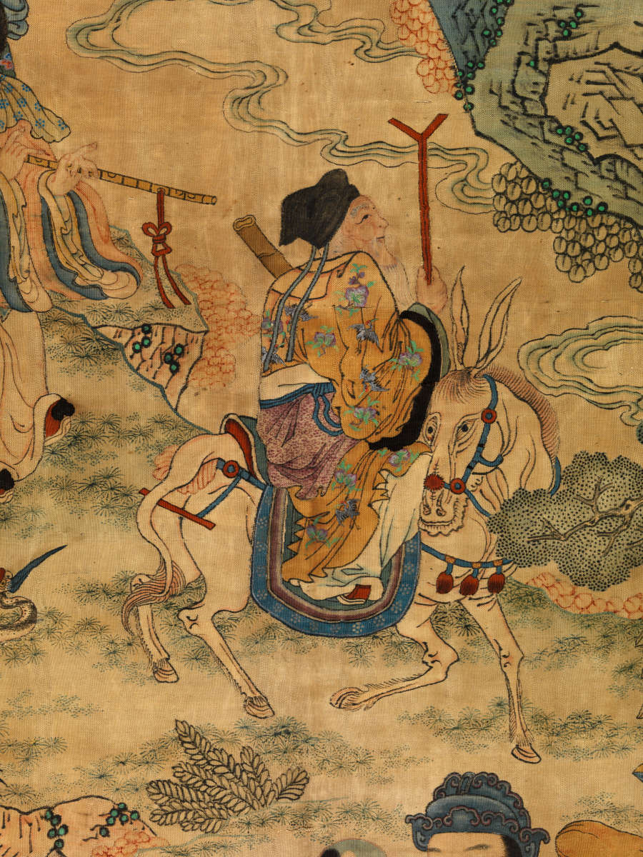 Detail of the scroll showing a robed figure with a long white beard holding a staff writing a white horse against a grassy and cloudy backdrop. Following them is a man playing the flute.