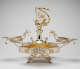 Elaborate silver double-oval-tray centerpiece with central fountain-like stem topper of winged-figurine holding up a suspended gold garland, surrounded by cherubs, and framed by swirling arms extending to two shallow dishes.