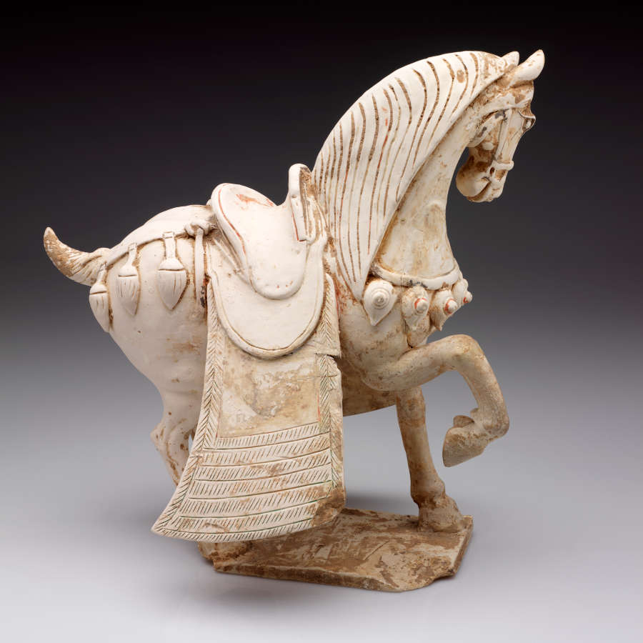 Another side-view of a white ceramic sculpture of a saddled, ornamented horse with one foot raised, a short wound tail and heavy mane. It stands on a thin connected base.