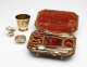 A traveling set with silverware. The traveling case has an orange colored fabric. There are multiple indents holding the silver, steel, and gilded cutlery and vessels.