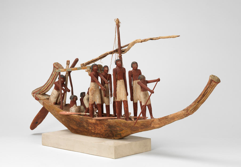 A carved wooden boat model with a sail tied on the mast manned by sailors with brown skin and white trousers holding oars and ropes.