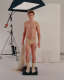 A fair-skinned male figure in beige underwear and socks stands on a black platform, holding draped fabric, and gazes into the camera in a studio setup with professional photography equipment. 