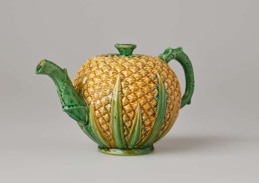 A teapot in the shape of a pineapple. Handle, foot, lid, and spout are green.