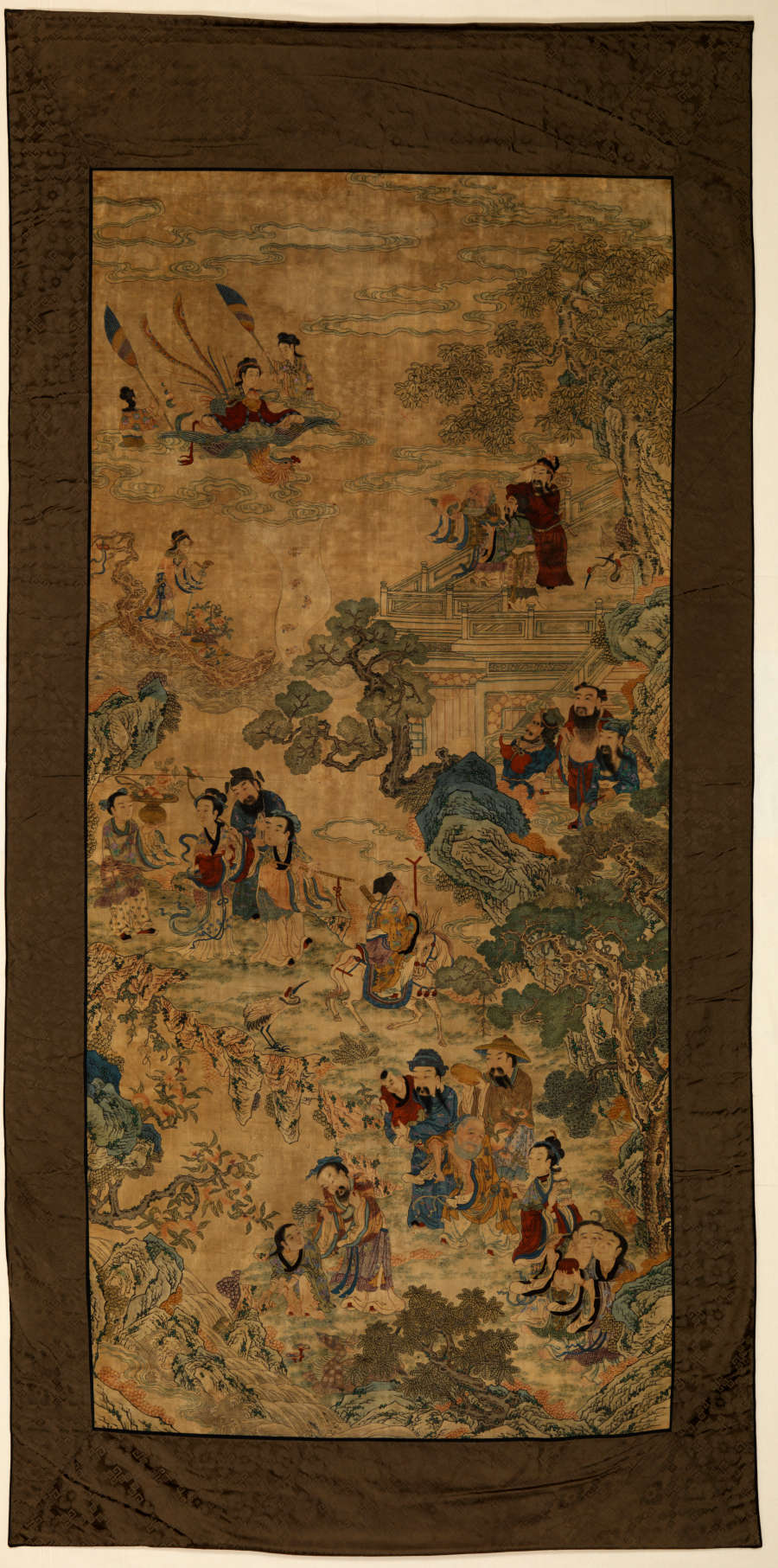 Vertically long rectangular scroll with a thick brown border. It features an illustration of several people engaging with another amongst trees, houses, and in boats on water.