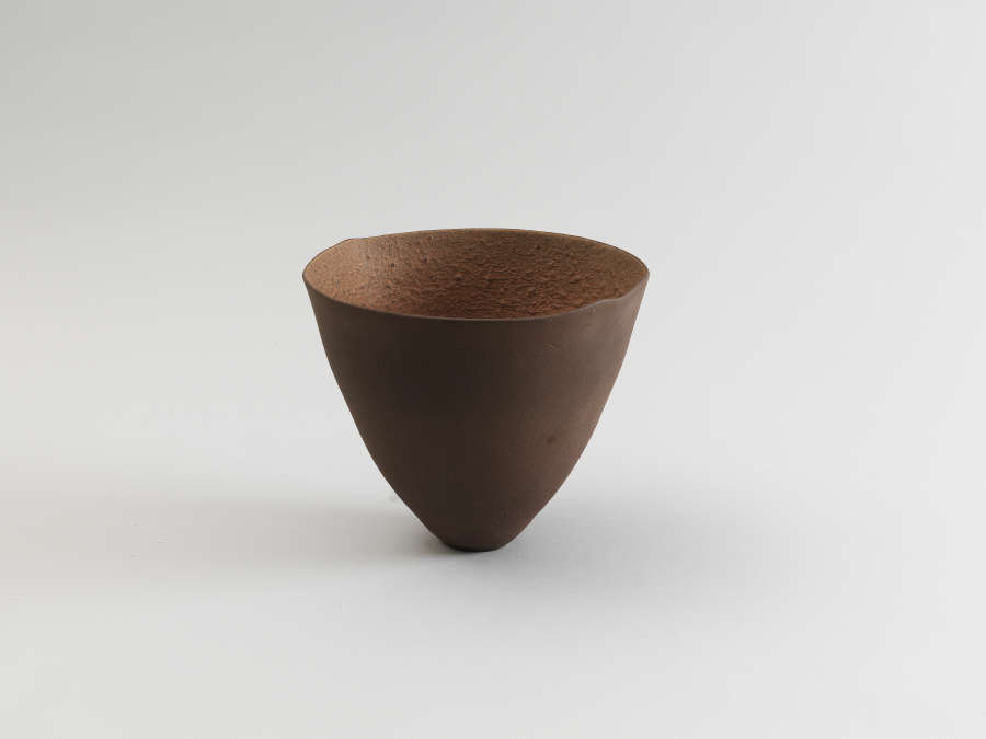 Dark brown ceramic bowl shaped like an inverted cone with a narrow, pointed base, featuring a rough, granular interior surface and a smooth, polished exterior.