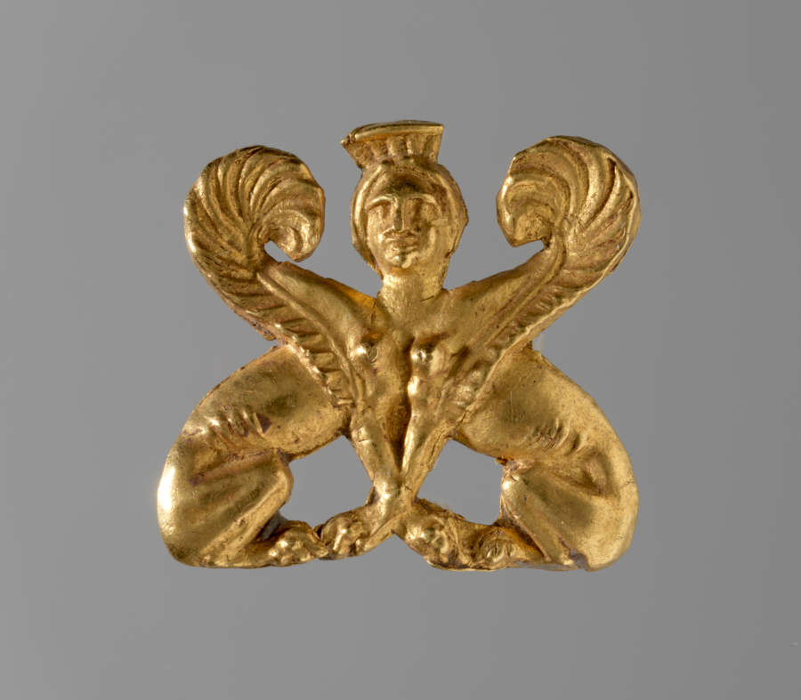 Gold ornament of two sphinx bodies facing one another. At their intersection is the gold crowned head of a woman, framed by two symmetrically arranged engraved wings.