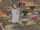 Ornate embroidery on a gray textile depicting an old monk in a white robe holding the handle of a golden object standing atop a backdrop of swirling clouds and flowers. 