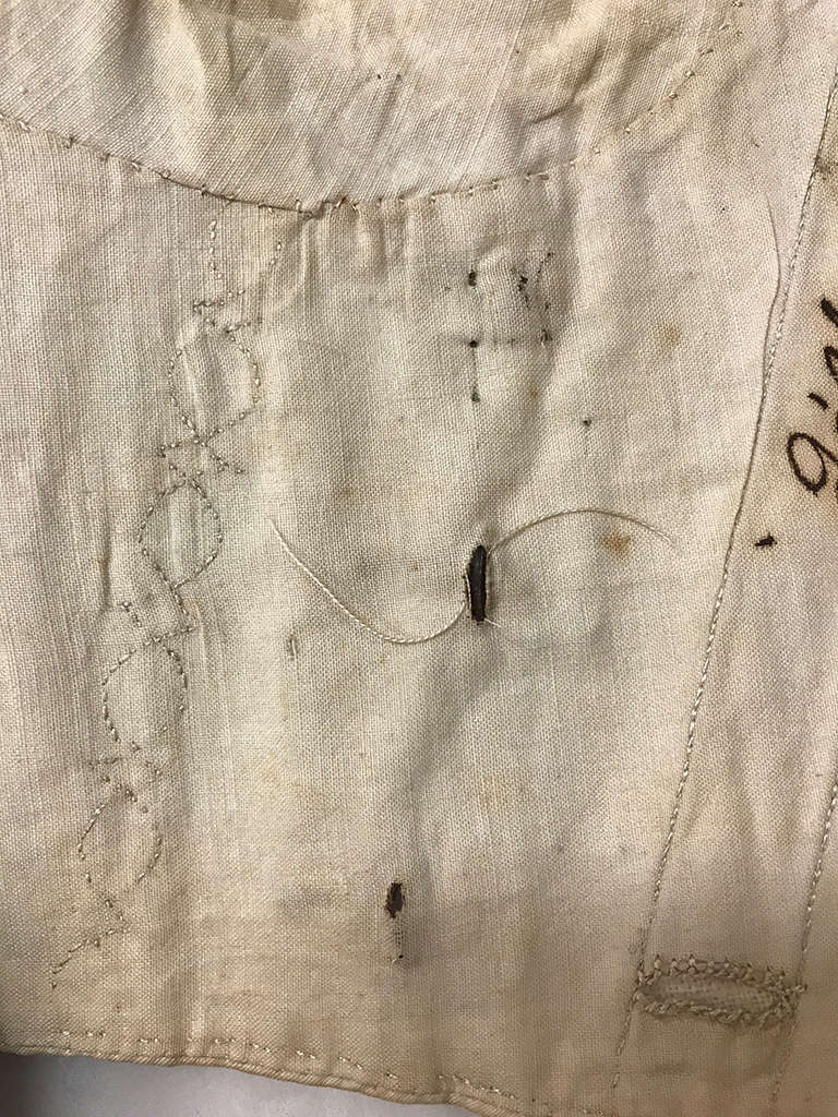 A close-up of an aged, cream-colored fabric with worn embroidery, small dark stains, and a loose thread protruding from a small hole. 