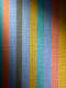 Close-up of painting surface, a spotlight illuminates evenly sized narrow vertical stripes in varied vivid colors, revealing pronounced raised canvas weave and small surface imperfections visible through the matte paint. 