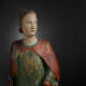 A weathered wooden statue of a figure with a solemn expression, with red and green drapery.