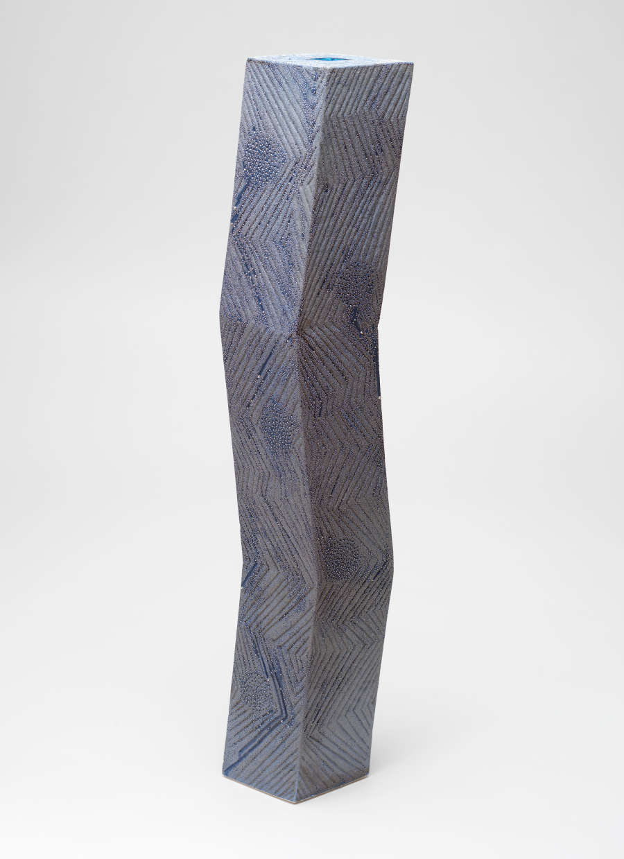 Tall, dark blue, chevron-patterned, rectangular column which bends angularly leftwards and rightwards. The irregular chevron patterning is formed by etchings overlaid by gray, powdery metallic material with a dew appearance.