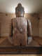 Rear view of a large wooden Buddha statue with visible cracks and signs of age, sat on a wooden platform in a warmly lit room.
