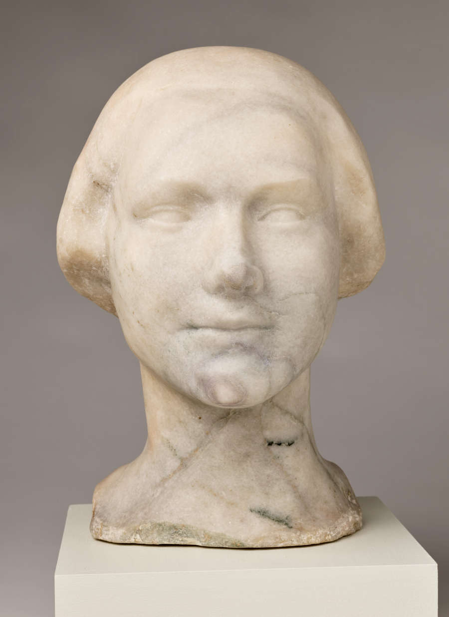 Front view of a blemished marble sculpture depicting the head of a female figure. The face features a faint smile and delicate details, framed by a short hairstyle.