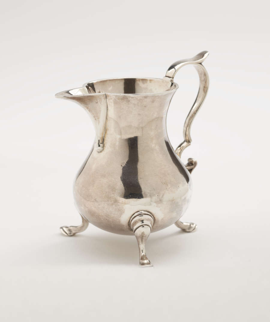 A silver creamer with a sculptural handle and three protruding feet.