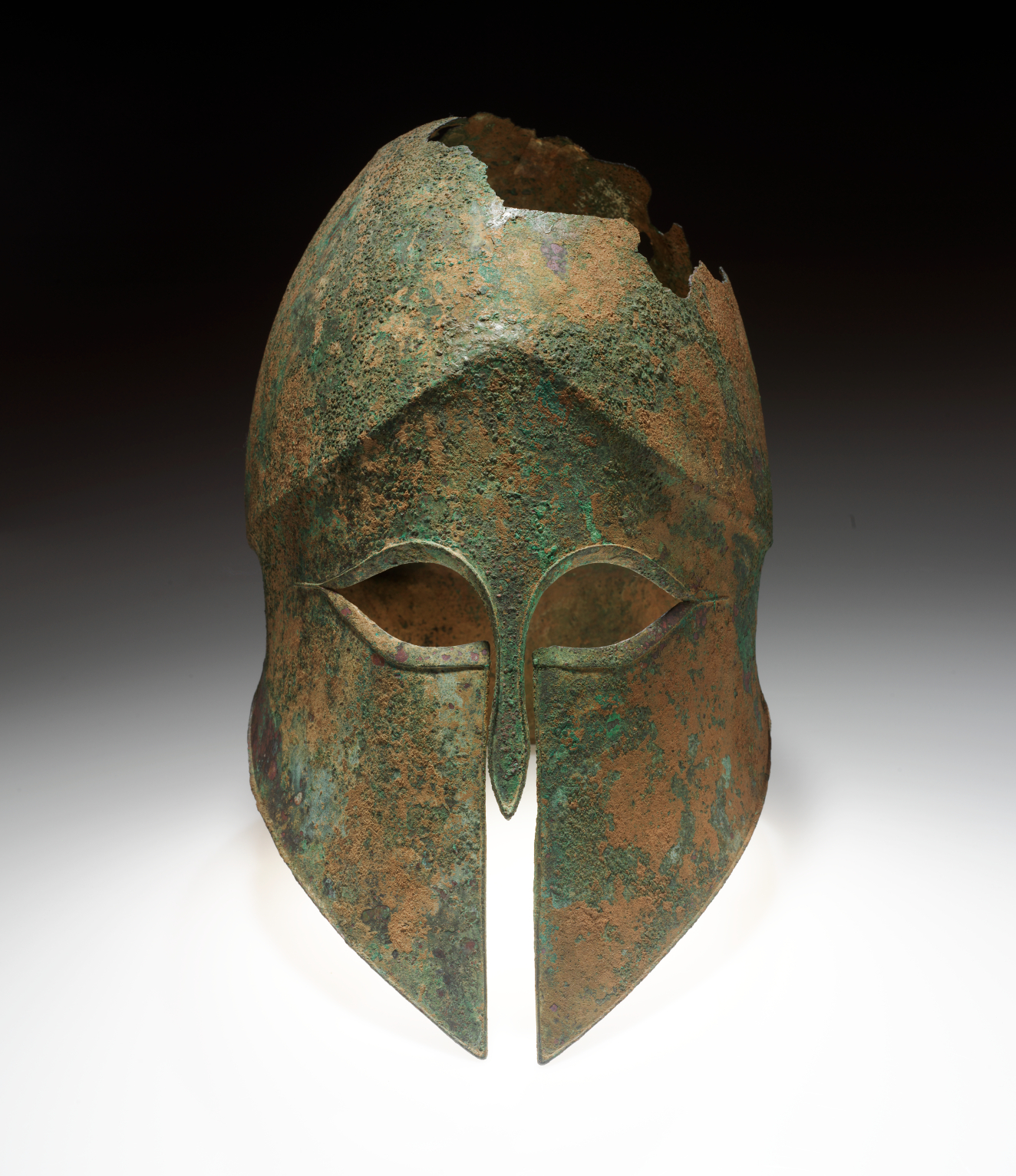 Bronze helmet with a narrow nose guard and sharp eye holes. The corroded surface is brown with significant green discoloration. The top has jagged sections missing along the crown.