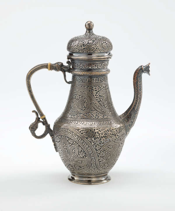 A silver coffee pot with detailed delicate decorations, handle with a lid hinged to it, and spout.