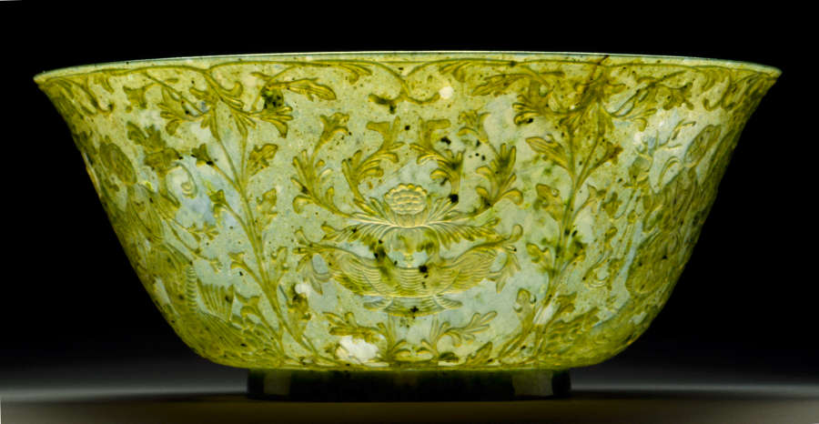 Jade bowl with sloped edges and a subtly flared mouth, with symmetrically arranged ornate floral etchings photographed in lighting making its surface appear bright white and yellow. 