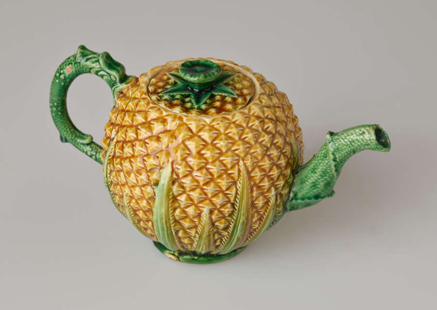 A teapot in the shape of a pineapple. Handle, foot, lid, and spout are green.