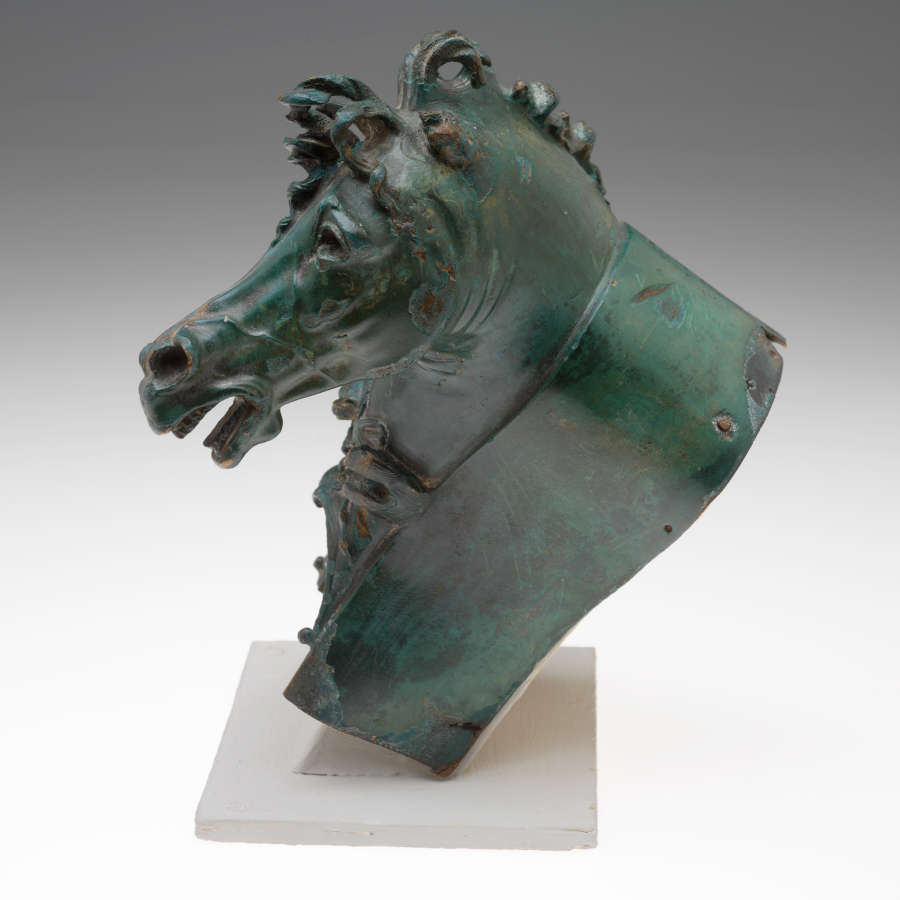 Side of a greenish-gray, patinated metal horse head ornament fitted onto a couch arm shaped pedestal.