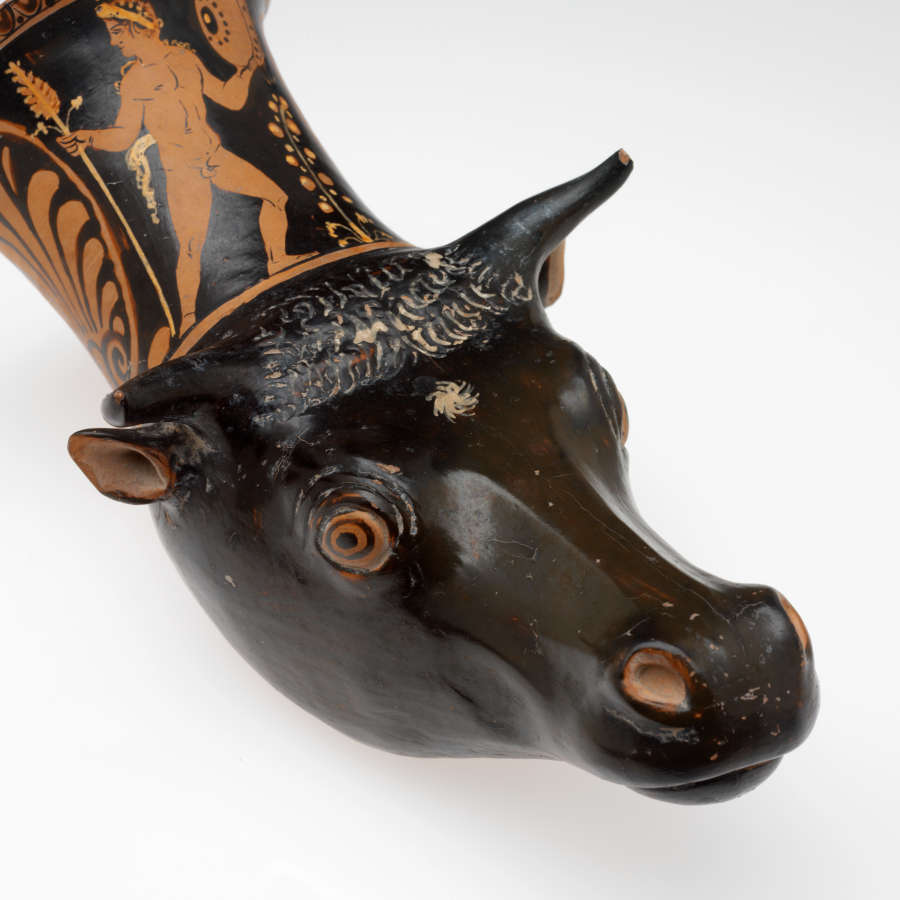 Black and terracotta cup with a sculpted cow’s head as its base, its top extending into the body of the cup, which is illustrated with men besides large floral motifs. 