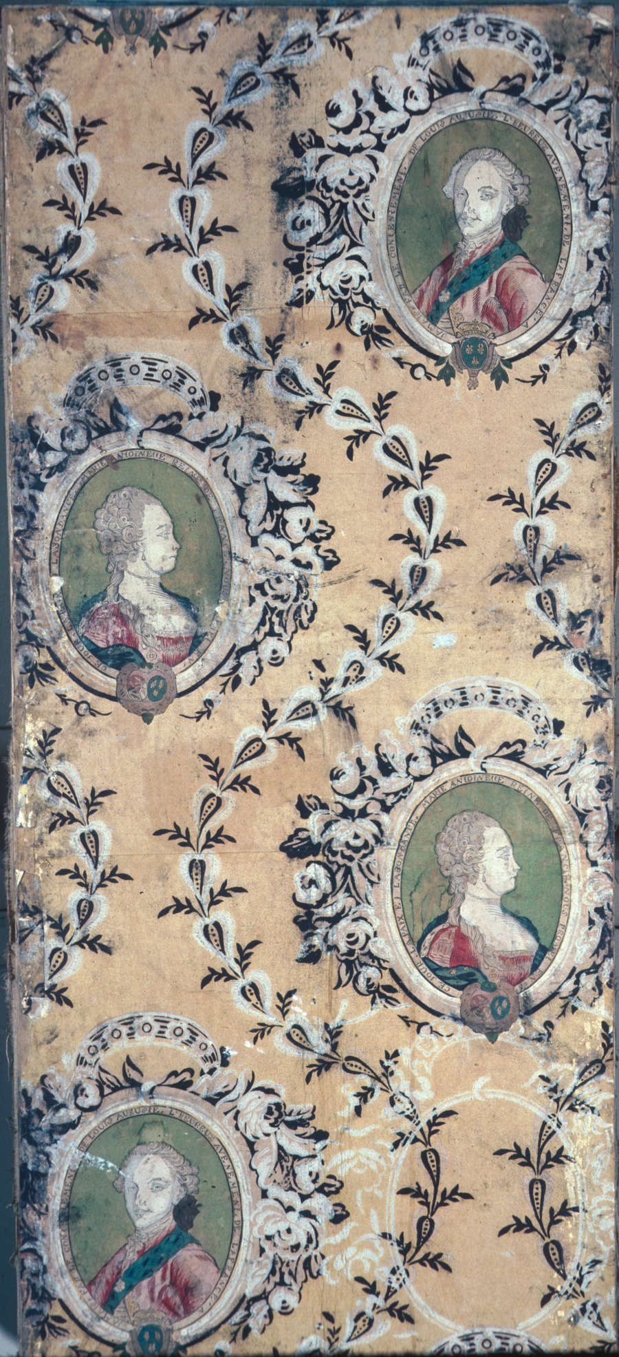 Tan, aged wallpaper of two repeating oval portraits, one of a man and the other a woman, both in elbarote dress encircled by delicate floral wreaths and leafy scrolls. 