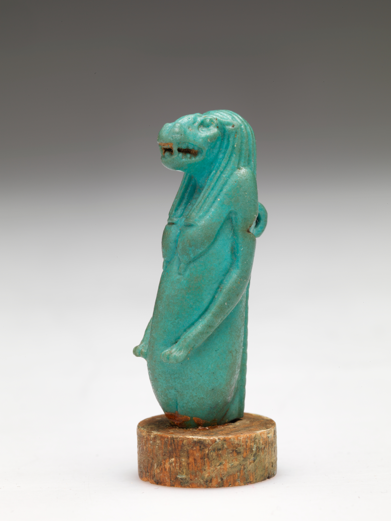 Angle profile of the aquamarine sculpture of a nude anthropomorphic hippo-headed woman with bare breasts, and long hair and arms. It is mounted onto a thick circular piece of stone.