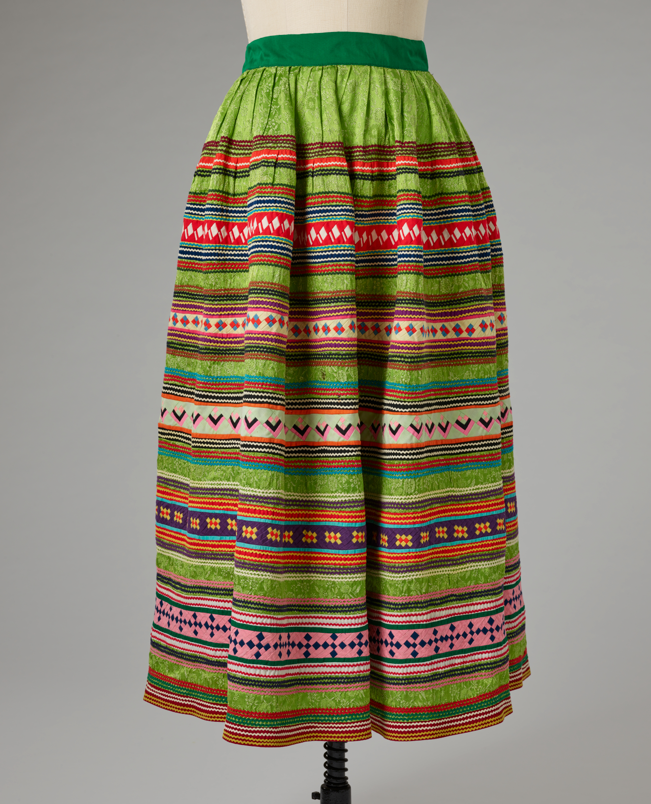 Green skirt on a mannequin, which is patterned with horizontal, warm and cool stripes, often containing patterns within them. The waistband is dark green. 