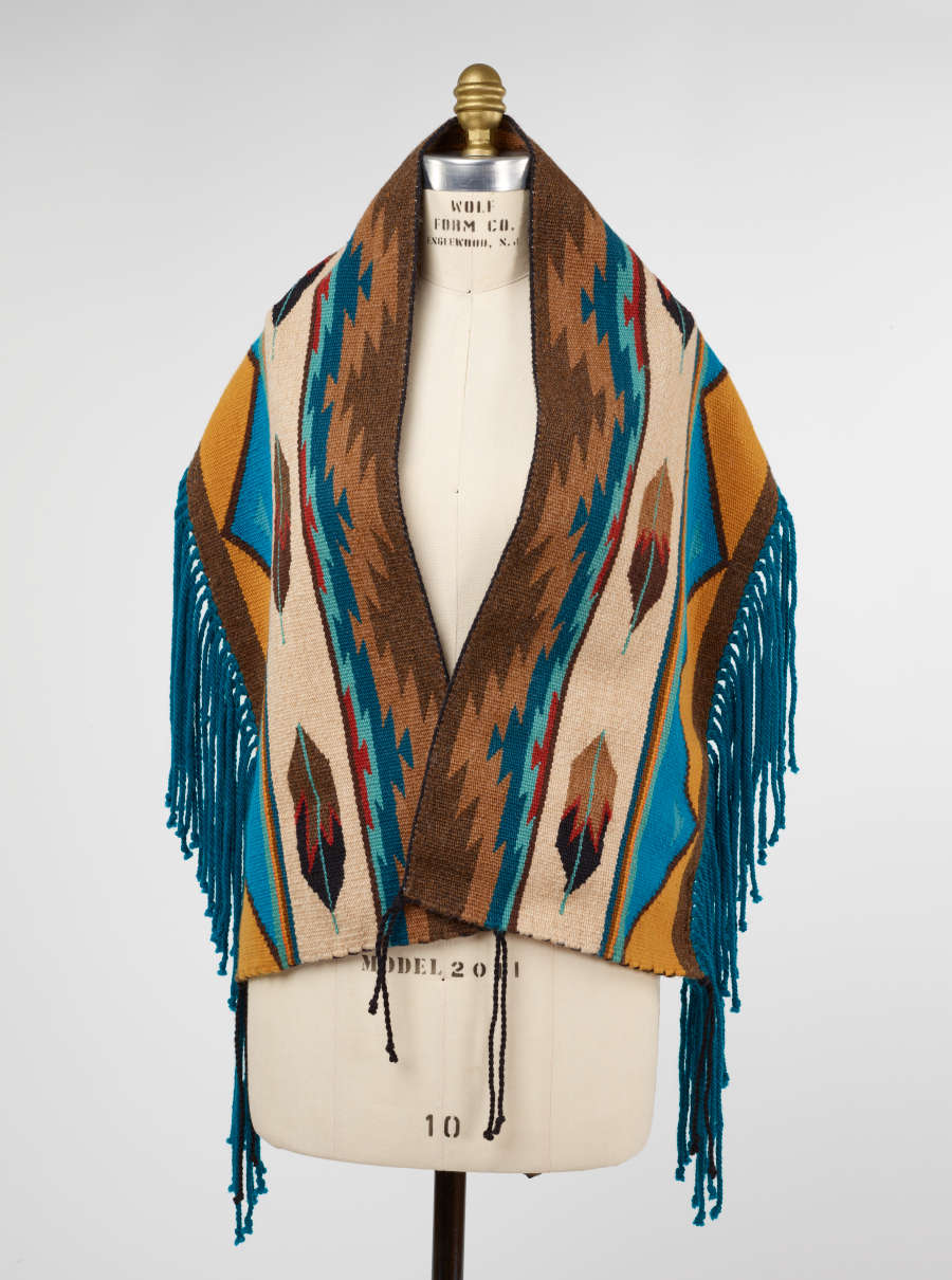 Multi-colored woven fabric with turquoise fringed ends and geometric patterning, draped around the torso of a mannequin. The fabric’s brown, blue, and red chevron pattern lines the neck and torso. 