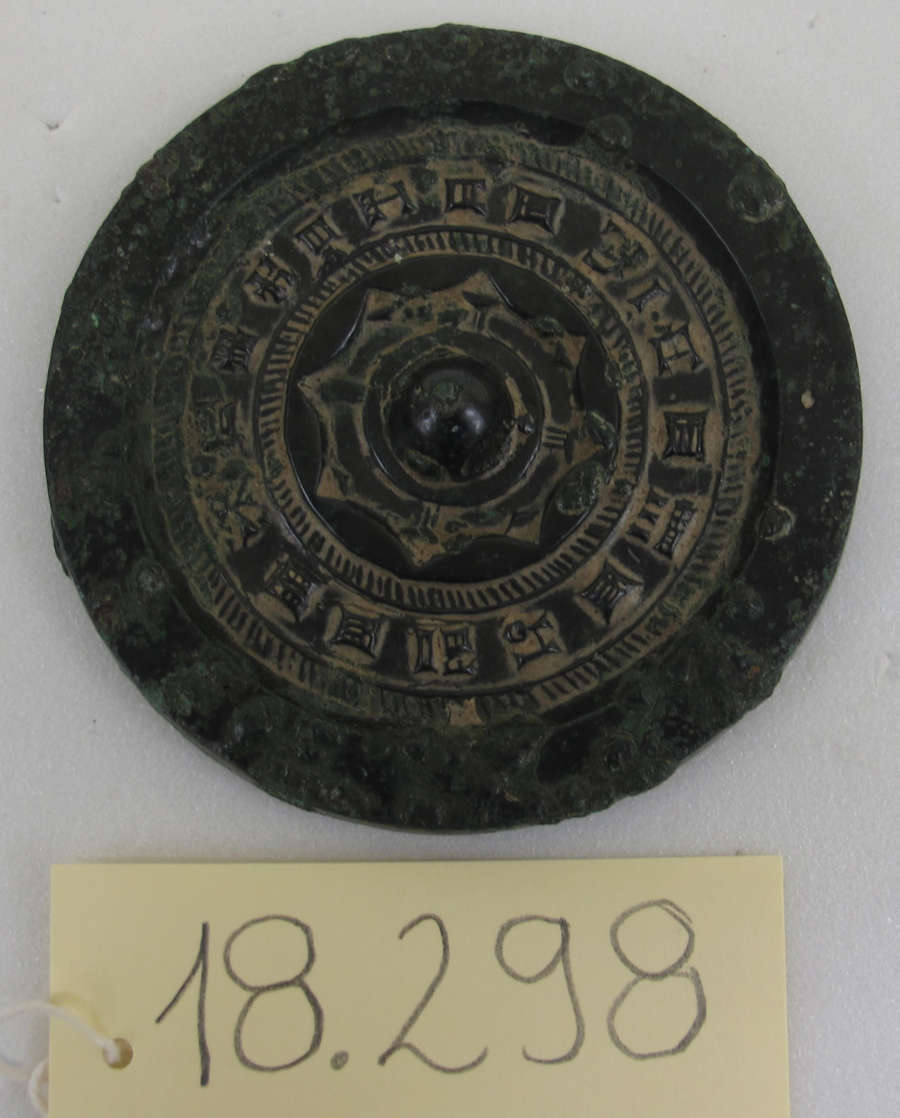 Back of a circular corroded bronze mirror in storage, embellished with a thick raised border, concentric engraved patterns, writing and a domed center. Below is a tag labeled ‘18.298’.