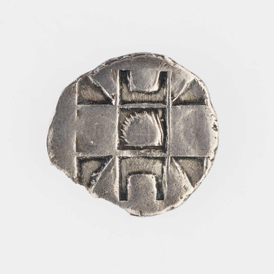 Silver coin with irregular edges. Embossed on its surface is a geometric pattern consisting of two pairs of intersecting lines dividing the coin into 9 regions.