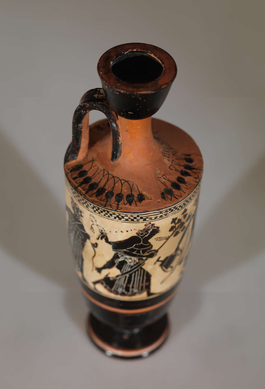 Angled top-view of a black and orange vase with illustrations of animals and figures. Highlighted is the vase’s thin orange fluted neck which has black floral motifs on its base.