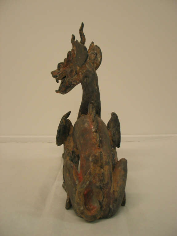 Back-view of an arched four-legged dragon with a lean body, open mouth, horned head, scaly back and a thin tail. The paint has worn into patches of browns and reds.