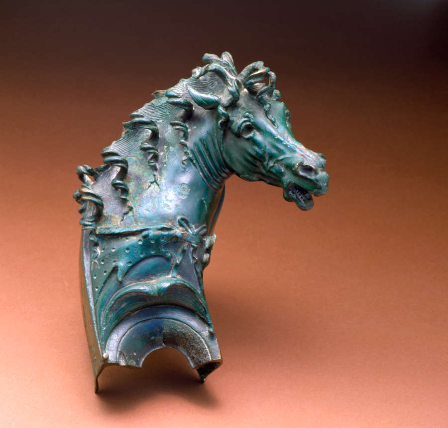 Greenish-gray metal ornament of a horse’s head, turned, with curls of hair falling along the ridge of its neck and its saddle. The horse’s mouth is partially open.