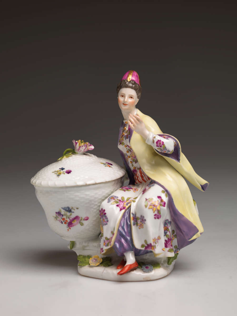 A sculptural figure sitting next to a large basket. The basket and figure are covered in floral decorations. The figure is wearing a hat, cream and purple clothing, and red shoes.